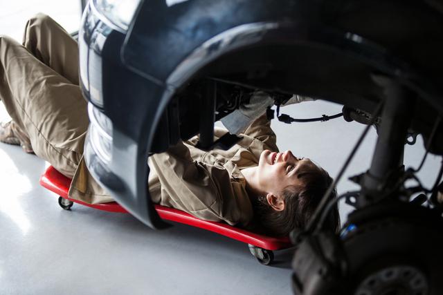 The image shows a female mechanic working underneath a car in a repair garage. She is lying on a red creeper while focusing on repairing parts of the vehicle. This photo can be used for content related to automotive repair services, promoting gender diversity in trades, car maintenance tips, or illustrating skilled labor in the automotive industry.