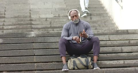 Senior african american man resting on urban stairs with bag water bottle and headphones