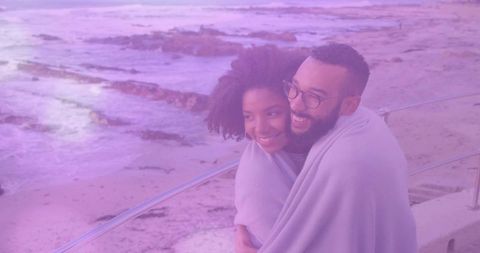 Couple embracing on seaside promenade wrapped in blanket enjoying windy sunset with purple tint