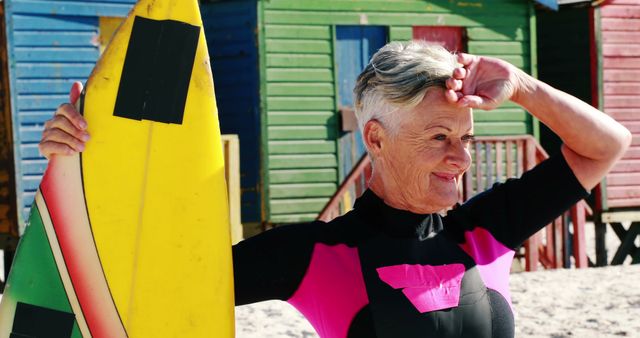 Elderly woman with short hair holding a yellow surfboard and smiling on a sunny beach. She wears a wetsuit with pink accents and stands in front of colorful beach huts. Ideal for promoting active senior lifestyles, retirement concepts, outdoor sports, and beach vacation themes.