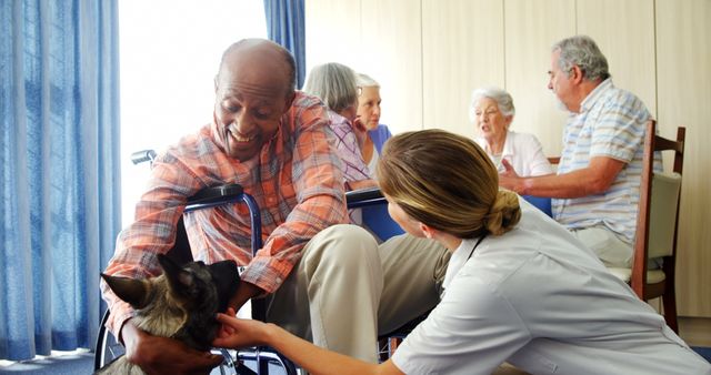 Senior man in wheelchair meeting therapy dog with assistance from a caregiver in a nursing home setting. Other elderly residents are engaged in background conversations, creating a warm and social atmosphere. This image showcases the benefits of animal therapy and the importance of social interactions for seniors. Ideal for use in articles about elder care, pet therapy, nursing homes, and senior health.