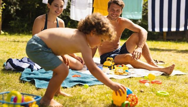 Family enjoying backyard picnic while child reaching for colorful toy wheel on sunlit lawn