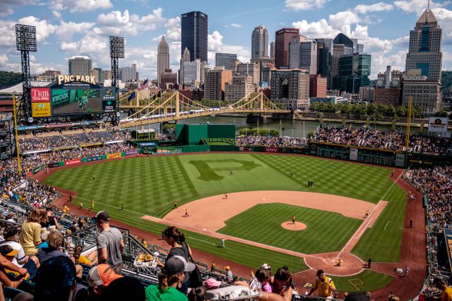 Baseball field on a sunny day with crowd enjoying a game, stadium filled with spectators. City skyline of Pittsburgh visible in the background. Ideal for content related to sports events, urban leisure, summer activities, tourism promotion, and city life.