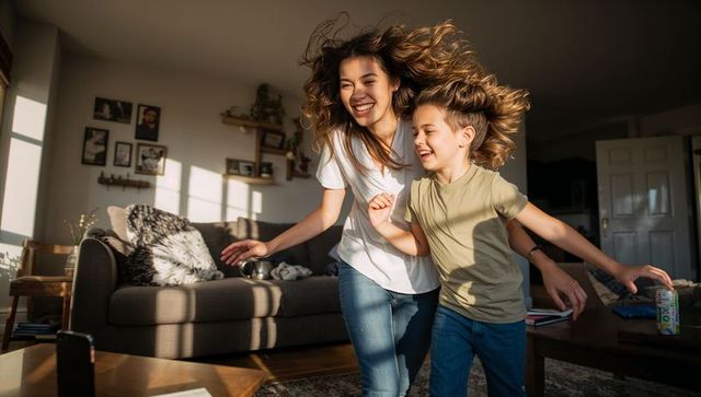 Smiling mother and son running and laughing in sunlit living room while recording on smartphone