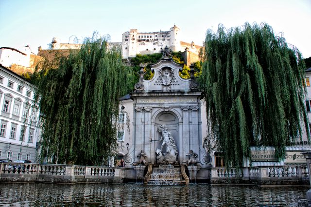 This picture shows a historic fountain in Salzburg, Austria with a fortified castle in the background, surrounded by lush greenery. The serene water and grand architecture make it ideal for travel and tourism marketing materials, cultural documentaries, or educational content related to European landmarks.