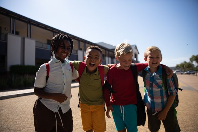 Four cheerful multiracial elementary schoolboys with arms around each other standing in front of a school building. They are smiling and appear to be enjoying their time together. This image is ideal for use in educational materials, advertisements promoting school diversity and inclusion, or any content related to childhood friendship and happiness.