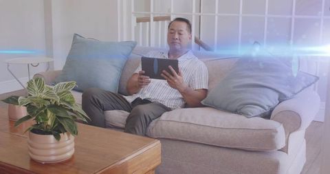 Chinese man relaxing on gray sofa using tablet in bright cozy living room