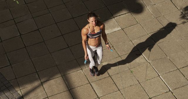 This image depicts an athletic woman engaging in a cardio workout by jumping rope outside on a sunny day. She is wearing sportswear and seems focused on her exercise routine. This photo can be used for articles or promotional materials related to fitness, healthy living, workouts, and outdoor activities. It is perfect for illustrating physical activity, motivation, and a healthy lifestyle.