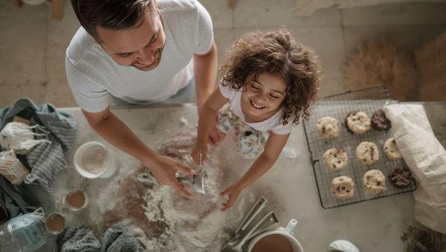 Father and daughter baking on flour-covered countertop, smiling, cookies cooling overhead