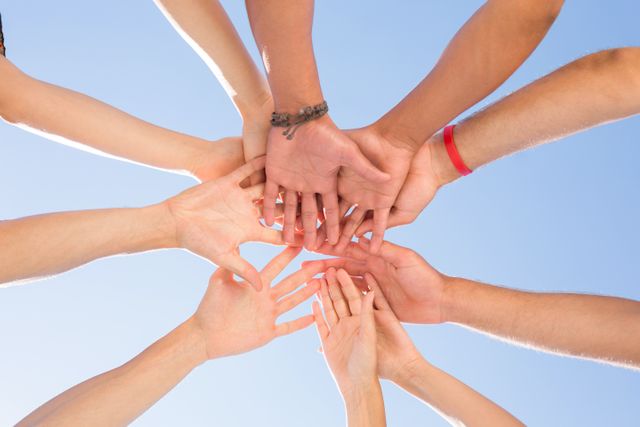 Friends stacking hands in a circle against a clear blue sky, symbolizing unity, teamwork, and support. Ideal for use in campaigns promoting teamwork, community building, and friendship. Perfect for illustrating concepts of collaboration, partnership, and solidarity in various contexts such as business, education, and social projects.