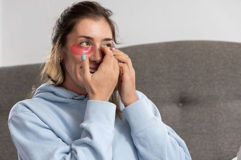 Young woman applying pink hydrogel under-eye patches on gray sofa for skincare self-care
