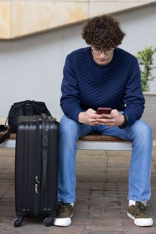 Young traveler sitting on bench checking phone with suitcase and backpack in urban plaza