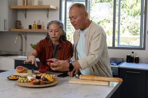 Mature couple arranging colorful fruit platter while slicing baguette on kitchen island