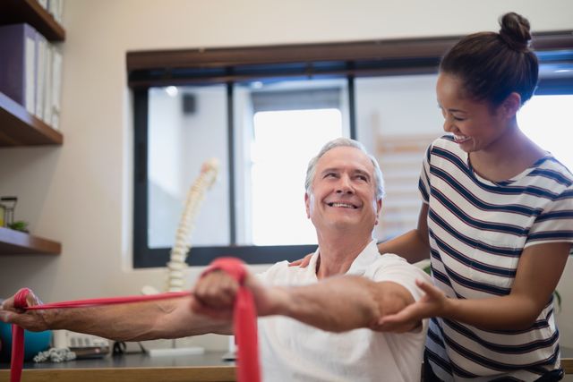 Smiling senior male patient pulling red resistance band while looking at female doctor - Download Free Stock Photos Pikwizard.com