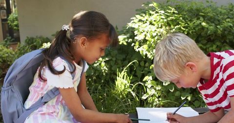 Two children studying outdoors on bench with backpack and notebook in sunlit garden