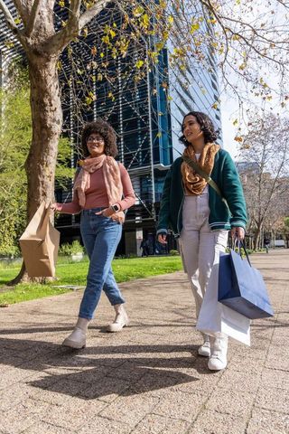 Friends walking through sunny urban park carrying shopping bags by glass office building