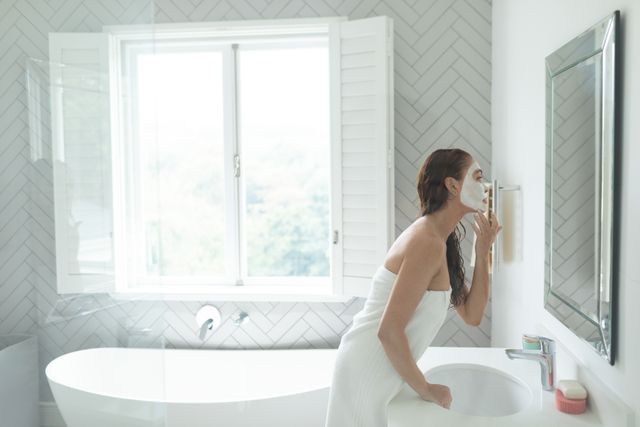 Woman wrapped in towel applying facial mask while looking in mirror in bright, modern bathroom. Ideal for content related to skincare routines, beauty tips, self-care practices, and wellness lifestyle. Can be used in blogs, advertisements, and social media posts promoting beauty products and home spa experiences.
