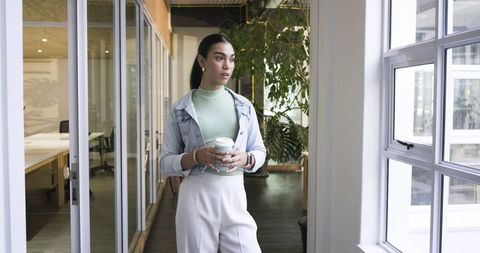 Asian professional woman standing by window holding takeaway coffee in coworking space