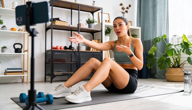 Recording female fitness trainer teaching live home workout on smartphone tripod, living room