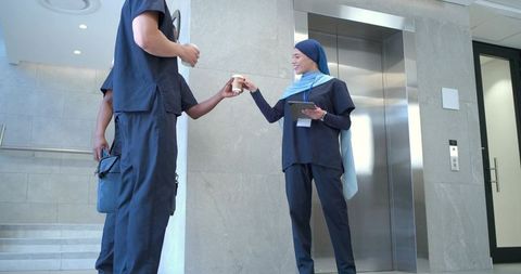 Healthcare team handing coffee in hospital lobby — nurse in hijab holding tablet