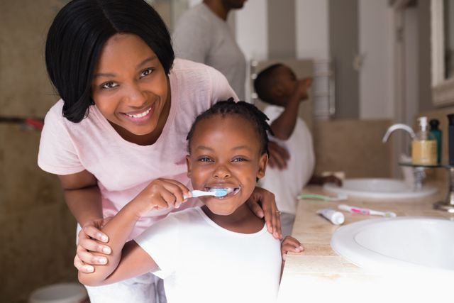 This image depicts a mother and her daughter engaging in a morning routine of brushing their teeth together in a bathroom. The mother is smiling and supporting her daughter, who is happily brushing her teeth. This photo can be used for promoting family bonding, dental hygiene, parenting tips, and health care products.