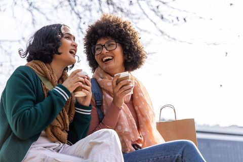 Sharing coffee and laughing together on urban bench, multicultural friends enjoying day