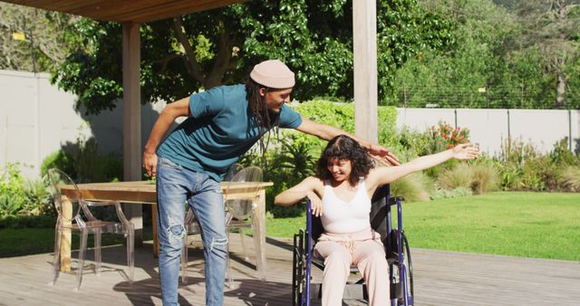 Happy biracial couple dancing on terrace in garden, woman sitting in wheelchair, man with dreadlocks. wellbeing and domestic lifestyle with physical disability.
