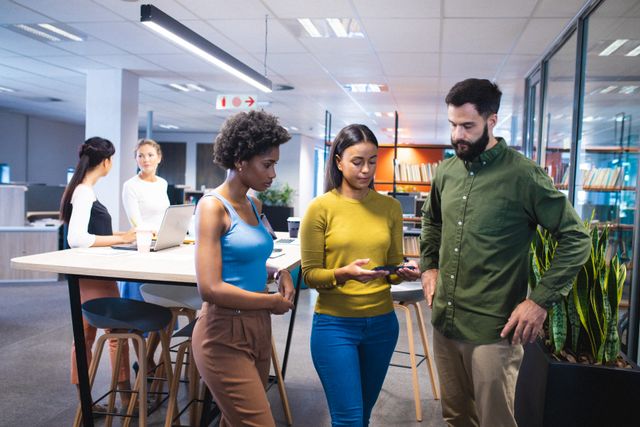 Multiracial business team standing in a modern office, discussing a project while looking at a smartphone. Ideal for use in articles about corporate culture, teamwork, modern office environments, and technology in the workplace.