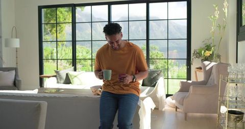 African man enjoying morning coffee while checking phone in bright modern bedroom