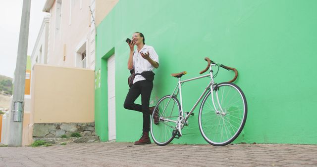 Front low angle view of a biracial man with long dreadlocks out and about in the city on a sunny day, standing on the stairs in the street, using a smartphone, with his bicycle leaning against the wall next to him in slow motion.