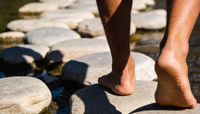 Barefoot man stepping across smooth river stones balancing on flat creek rocks sunlit