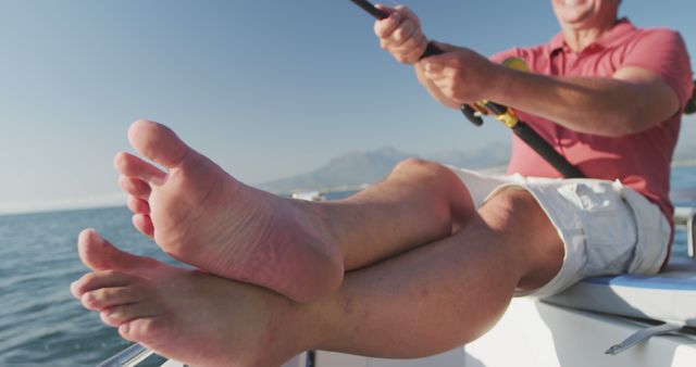 Low section of caucasian man sitting barefoot and fishing from deck of small boat on a sunny day. Leisure, hobbies, free time, family, travel and vacations.