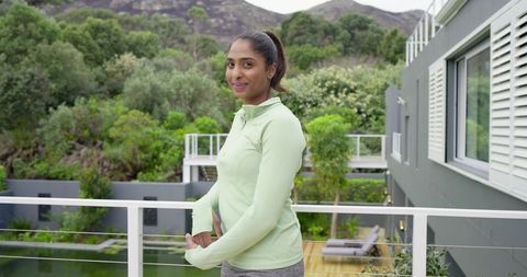 Indian woman wearing activewear standing on modern balcony overlooking pool and garden