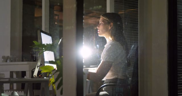 Young businesswoman focused while working late at her desk in a modern office, illuminated by computer screen and desk lamp. Use for themes related to work dedication, night shifts, corporate environments, and professional women in business settings.