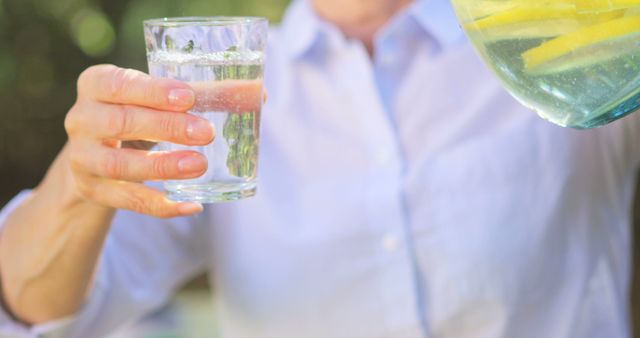 Person holding clear glass of lemon water with lemon slices visible in sunny outdoor environment. Ideal for health and wellness blogs, summer refreshment ads, or healthy lifestyle promotions.