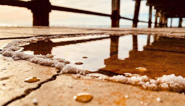 Golden-hour reflection in shallow puddle revealing seafoam and pier column silhouettes