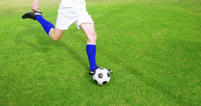 Soccer player in white uniform and blue socks kicking ball on green field. Perfect for advertisements related to sports equipment, fitness campaigns, teamwork promotion, athletic events.