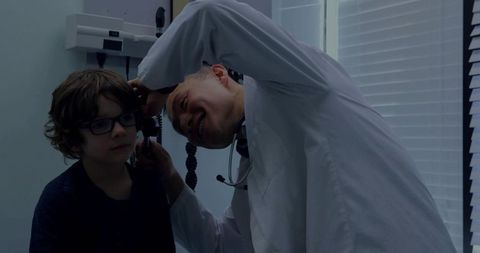 Asian pediatrician examining young boy's ear with otoscope in clinic exam room