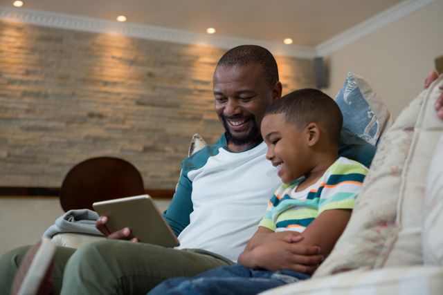 Father and son sitting on a comfortable sofa in a cozy living room, both smiling while looking at a digital tablet. Ideal for use in articles or advertisements about family bonding, parenting, technology in family life, or home leisure activities.