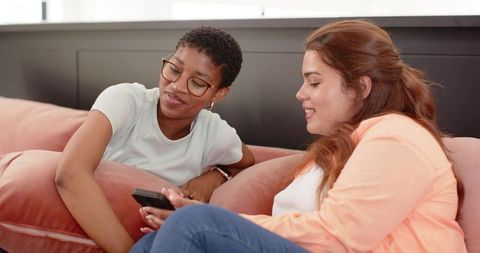 Two friends relaxing on pink sofa sharing smartphone, laughing and chatting in cozy living room