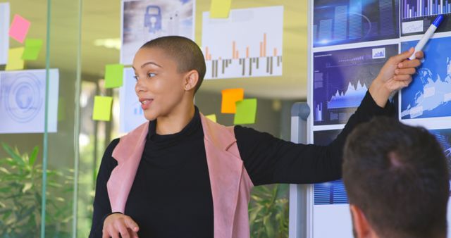 Businesswoman conducting presentation with data charts in modern office with glass walls and multiple sticky notes. Useful for workplace productivity, business strategy, and corporate communication themes.