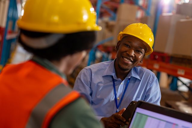 Smiling African American man engaging in conversation with female coworker using laptop in warehouse. Both wearing hard hats, indicating a focus on safety. Ideal for illustrating teamwork, logistics, and warehouse operations in industrial settings.