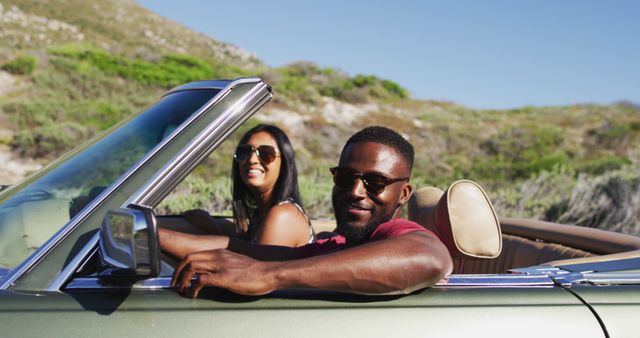 Portrait of african american couple waving and having fun while sitting in the convertible car on road. road trip travel and adventure concept