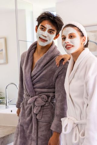 Asian man and african american woman wearing face masks and bathrobes enjoying at-home spa
