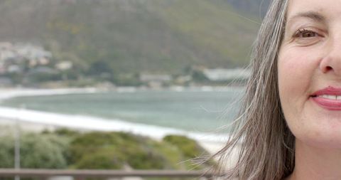 Mature woman smiling on balcony overlooking tranquil coastal bay with soft-focus seascape