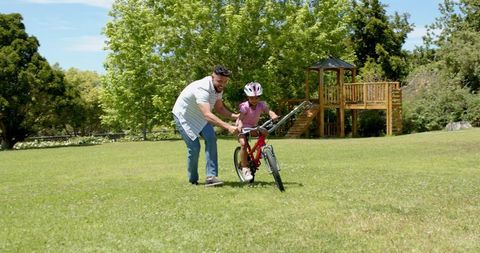 Father Helping Daughter Ride Bicycle in Park Under Blue Sky