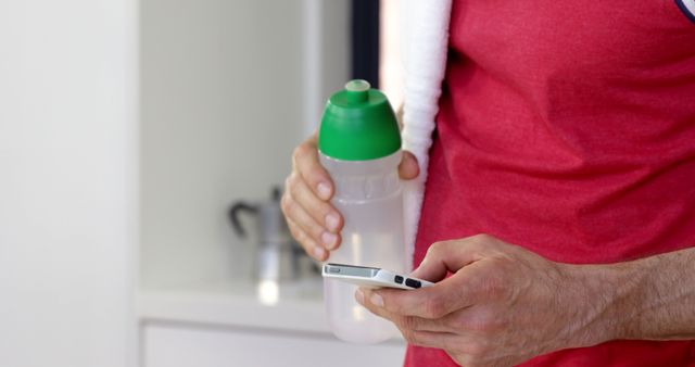 A Caucasian middle-aged man is holding a water bottle and checking a smartphone, with copy space. He appears to be taking a break from physical activity, reviewing his workout progress or staying connected.