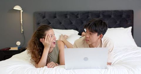 Diverse young couple lying on bed using laptop, sharing relaxed moments in modern cozy bedroom
