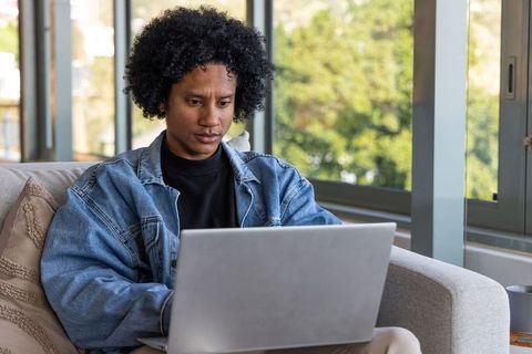 African american man working from home on laptop in cozy living room wearing denim jacket