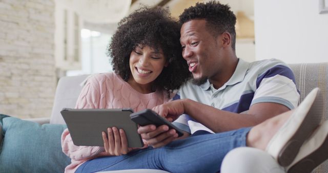 A couple is smiling while using a tablet and smartphone together, sitting on a cozy couch at home. Ideal for use in advertising tech gadgets, portraying happy relationships, showcasing modern home lifestyles, and promoting digital communication and leisure activities.